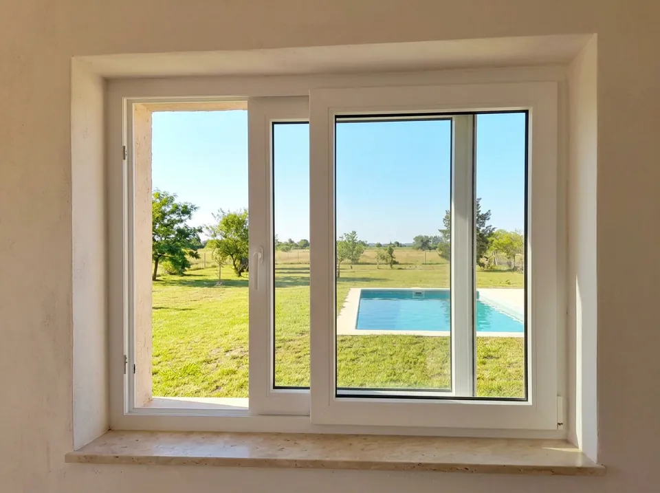 Ventana corrediza de PVC blanco, entreabierta, con el jardín y la pileta afuera